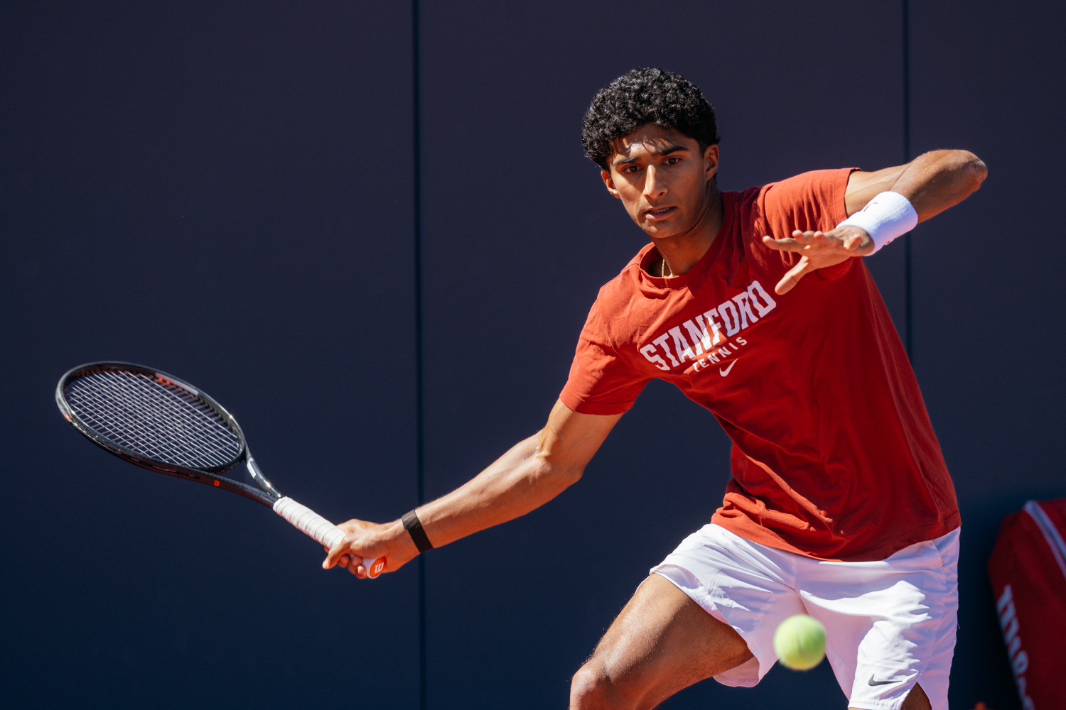 Stanford tennis player Samir Banerjee in action, wearing a red shirt with "Stanford Tennis" visible.