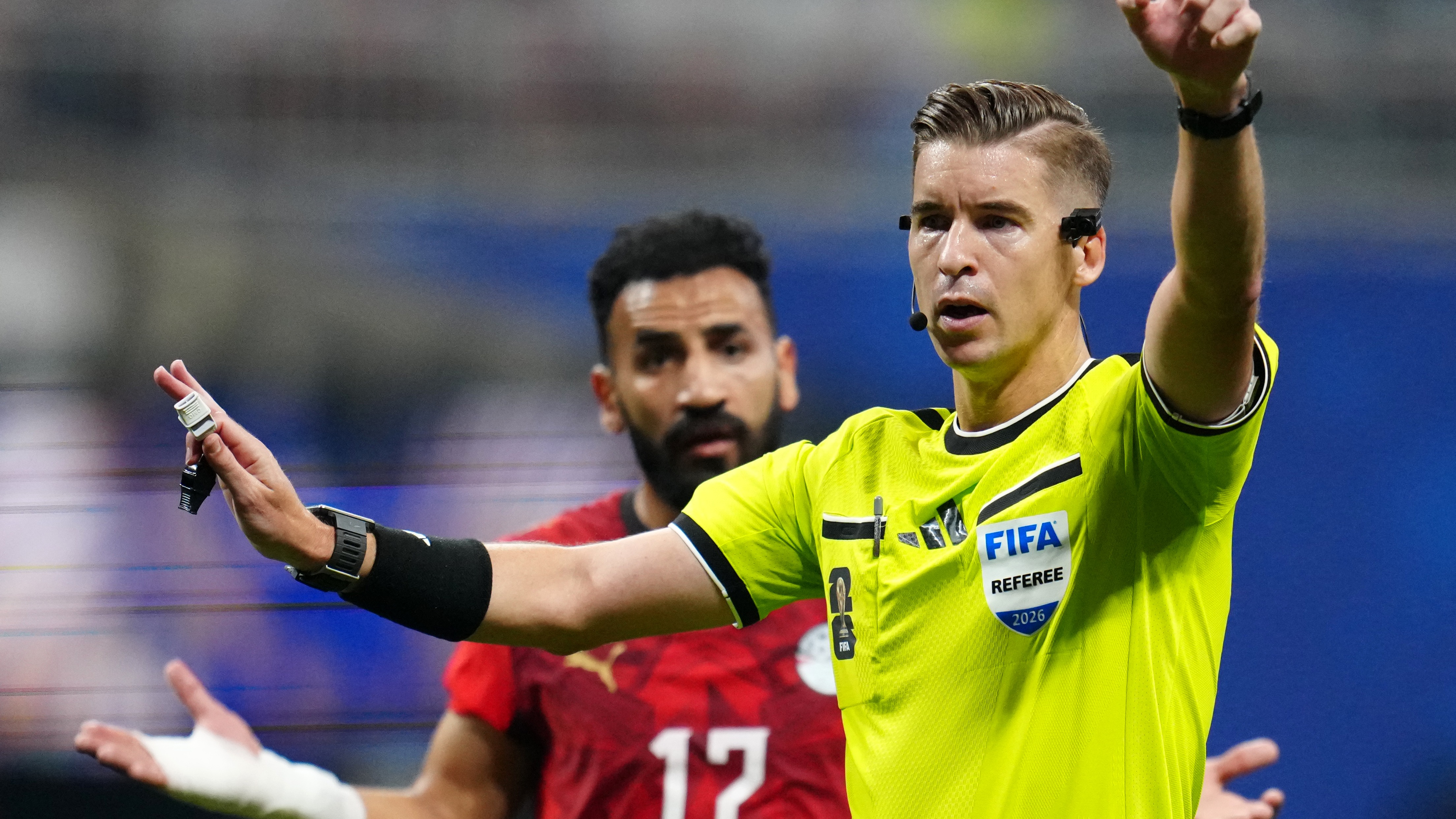 FIFA referee gesturing during a soccer match, player in red in background