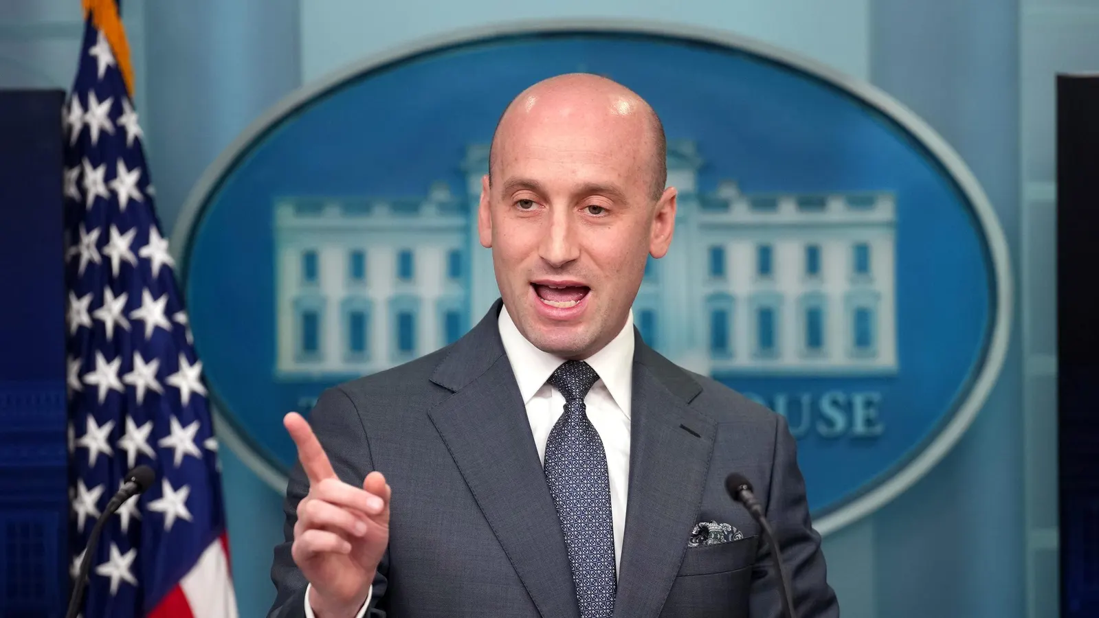Man speaks at podium with US flag and White House seal, discussing H-1B visa.